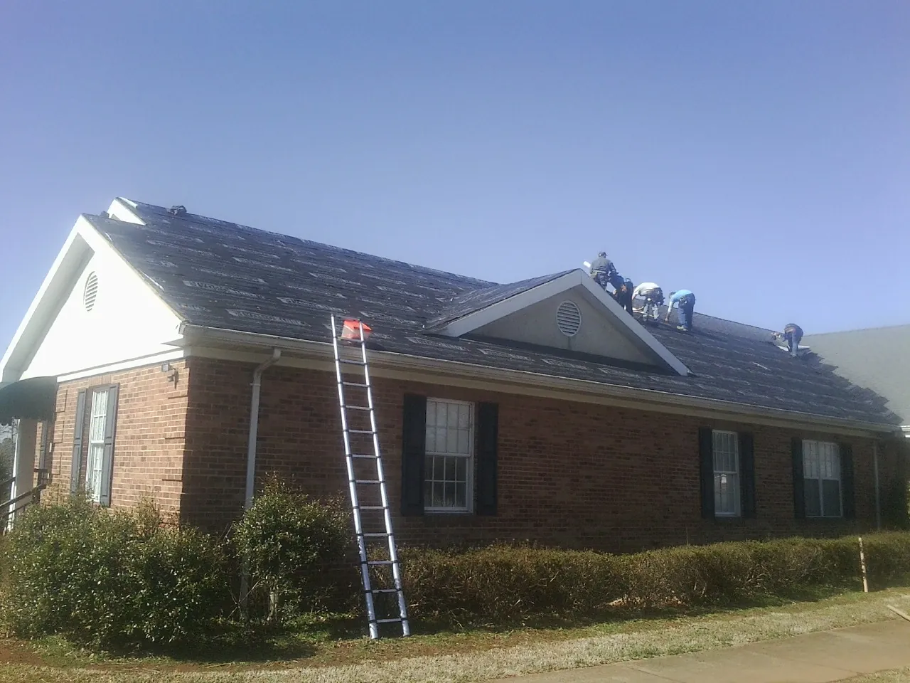 Crew tearing shingles off a brick ranch roof with a ladder set — Emergency Roof Repair work by our crew in Cambria Heights
