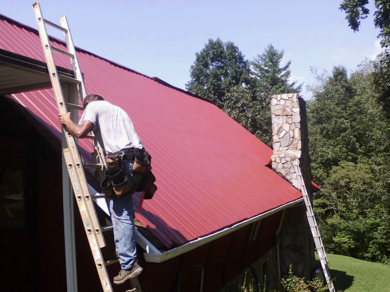 Roofer on a ladder beside a red metal roof and stone chimney — Soffit & Fascia Repair by Roofing Brothers in Cambria Heights